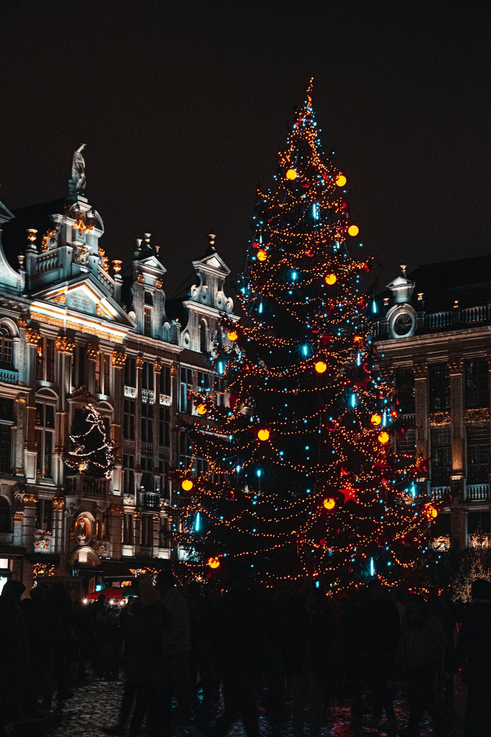 Albero di Natale con luci arancioni e blu in una piazza di una città europea.